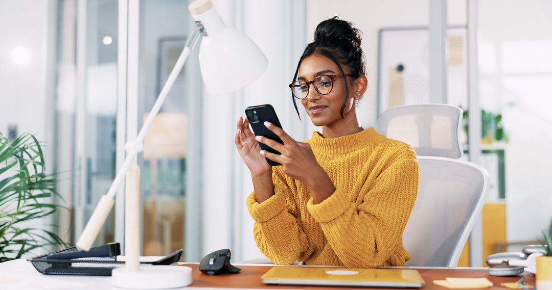 Woman in a yellow sweater sitting at a desk, smiling while looking at her phone in a modern office